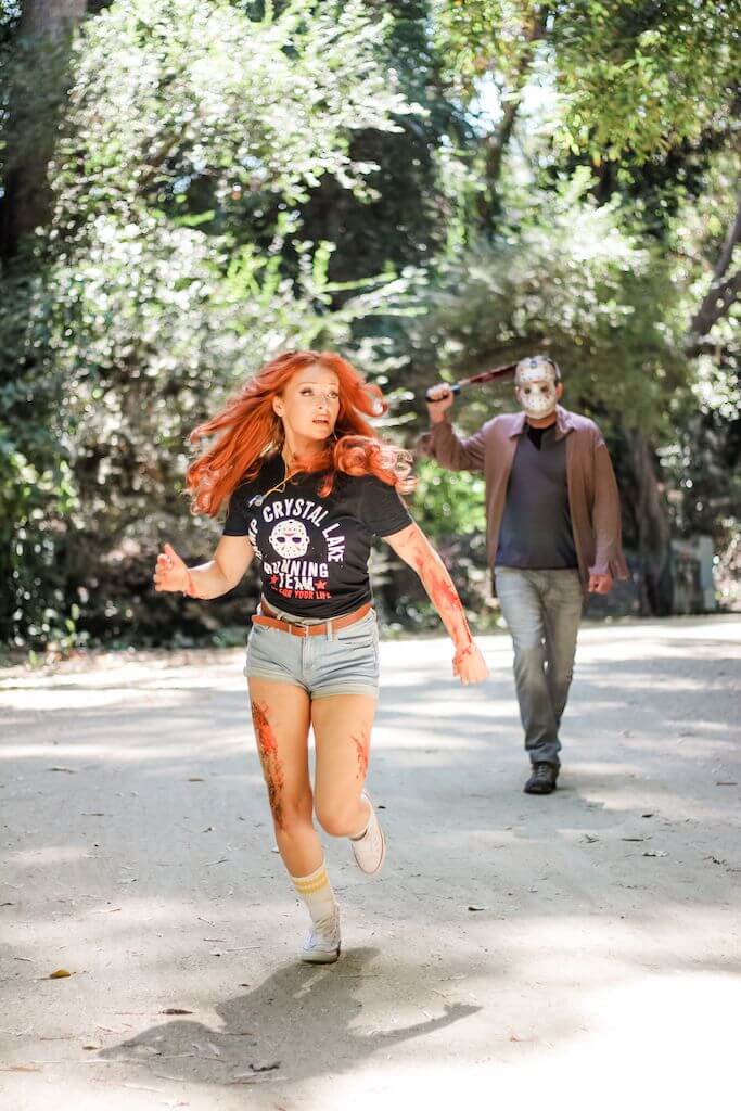 There is a camp counselor with red hair, a black camp crystal lake running team shirt and denim shorts running, while Jason Voorhees stands behind her in a hockey mask and brown shirt, while holding an axe.