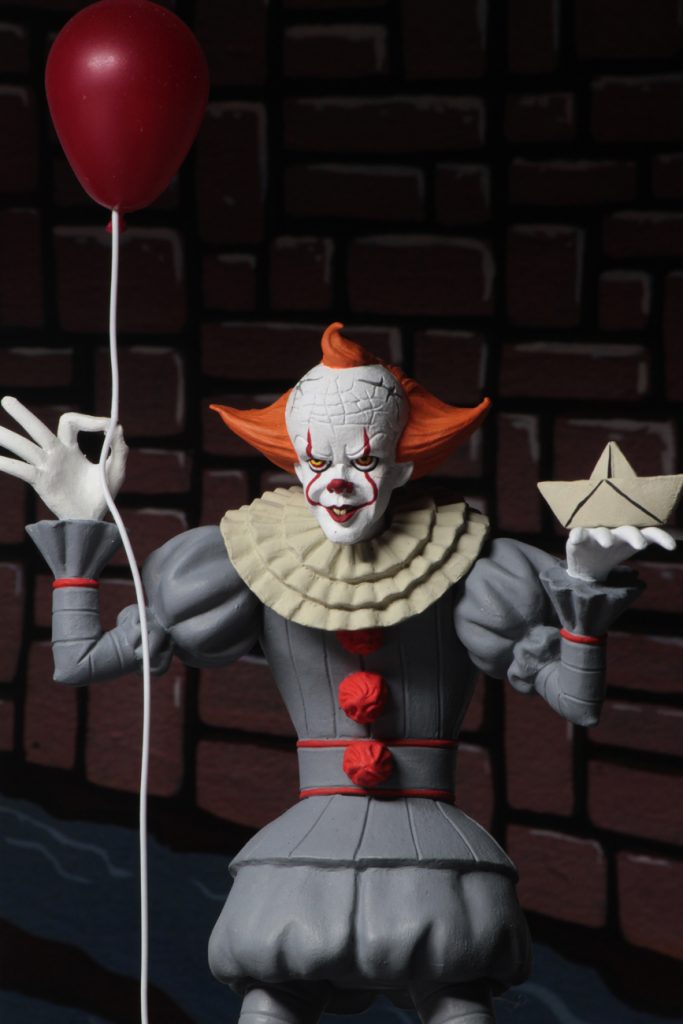 This is a NECA Toony Terrors It 2017 Pennywise action figure and he is wearing a grey clown suit that has three red balls and a white collar, with white gloves, balls on his shoes, a white face and orange hair and is holding a white boat and red balloon.